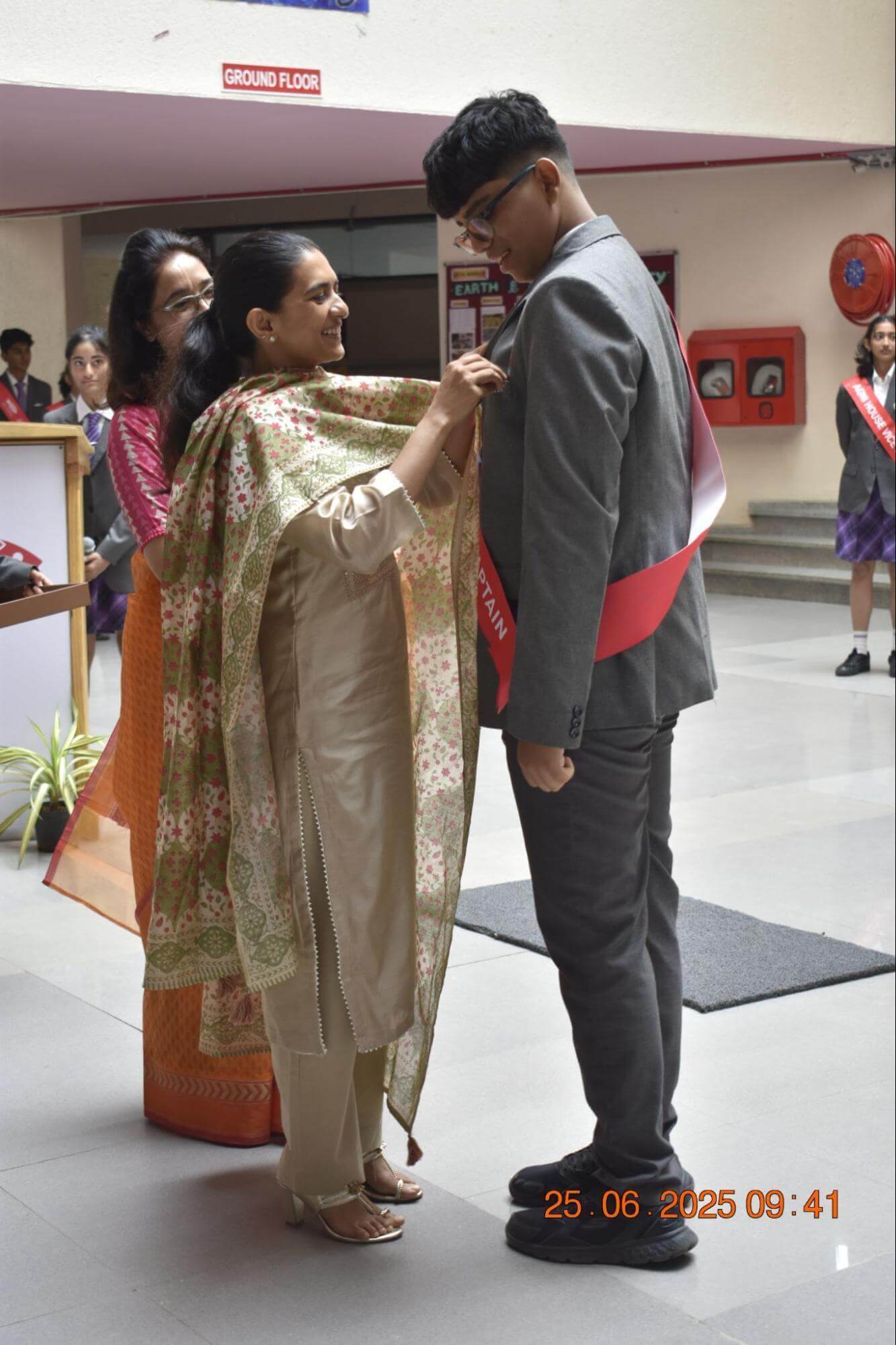 A woman pins a badge onto a student wearing a “CAPTAIN” sash during an Investiture Ceremony at Ekya School. Other students in uniforms stand in the background.